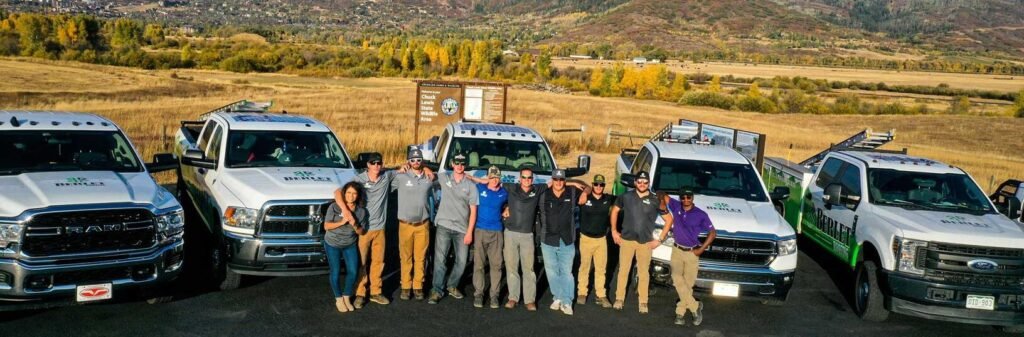 Professional roofing team posing outdoors with company vehicles against a mountain backdrop.